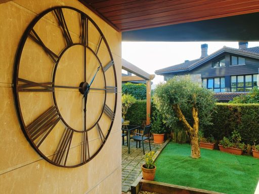 Reloj de pared grande en un jardín con césped y plantas decorativas.