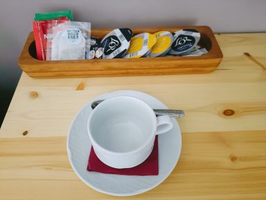 Servicio de café e infusiones Taza blanca sobre un plato con un servilleta roja y un recipiente de madera con sobres y tés.